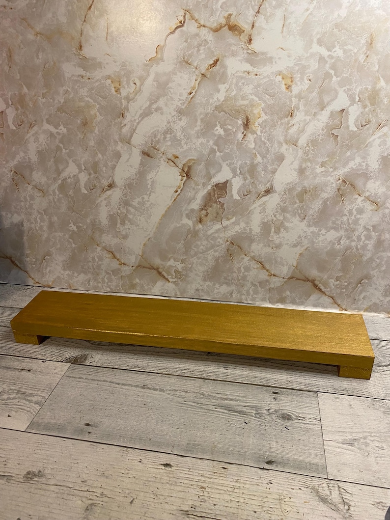 May include: A rectangular wooden riser with a smooth, metallic gold finish. The platform is elevated by two short, block-style feet on either end, creating a low-profile stand suitable for displaying various items on a flat surface.