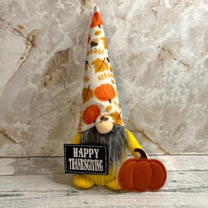 May include: A decorative gnome figurine featuring a tall, pointed hat with an orange and yellow autumn leaf and pumpkin pattern. The gnome has a long gray beard and holds a black rectangular sign that reads HAPPY THANKSGIVING in white.