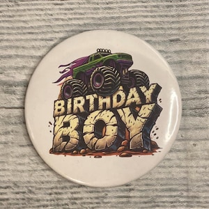 May include: A white button with a cartoon illustration of a green and purple monster truck on a rocky surface. The text "BIRTHDAY BOY" is in large, textured letters below the truck. The button is round and has a glossy finish.