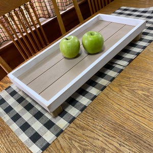 May include: A white wooden tray with a light brown wood top and four wooden legs. The tray is sitting on a black and white checkered placemat. Two green apples are on the tray.
