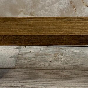 K&ouml;nnte beinhalten: Ein rechteckiges Holzregal mit einer mittelbraunen Beize und sichtbarer horizontaler Holzmaserung. Das Regal hat eine glatte, flache Oberfl&auml;che und gerade Kanten, die die nat&uuml;rliche Textur und Farbe des Holzes deutlich zur Geltung bringen.