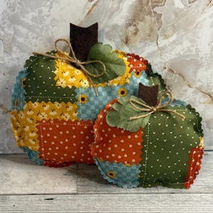May include: Two fabric pumpkins featuring a patchwork design of green, yellow, and orange floral and checkered patterns. Each pumpkin has a dark brown felt stem, a green fabric leaf, and a decorative jute twine bow tied around the stem.