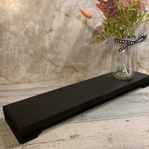 May include: A rectangular black wooden riser with a flat top and short feet. A clear glass mason jar sits on the right side of the riser, containing a bouquet of pink and green dried flowers tied with a black and white checkered ribbon.