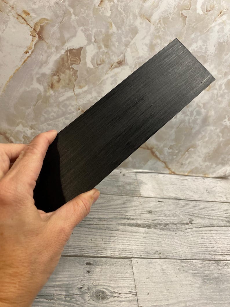 K&ouml;nnte beinhalten: Ein rechteckiger Block aus dunkel anthrazitfarbenem Holz mit einer sichtbaren, fein gemaserten Textur, die horizontal &uuml;ber die Oberfl&auml;che verl&auml;uft. Das Objekt ist massiv, flach und weist scharfe, saubere Kanten auf, was ein einheitliches Erscheinungsbild ergibt.