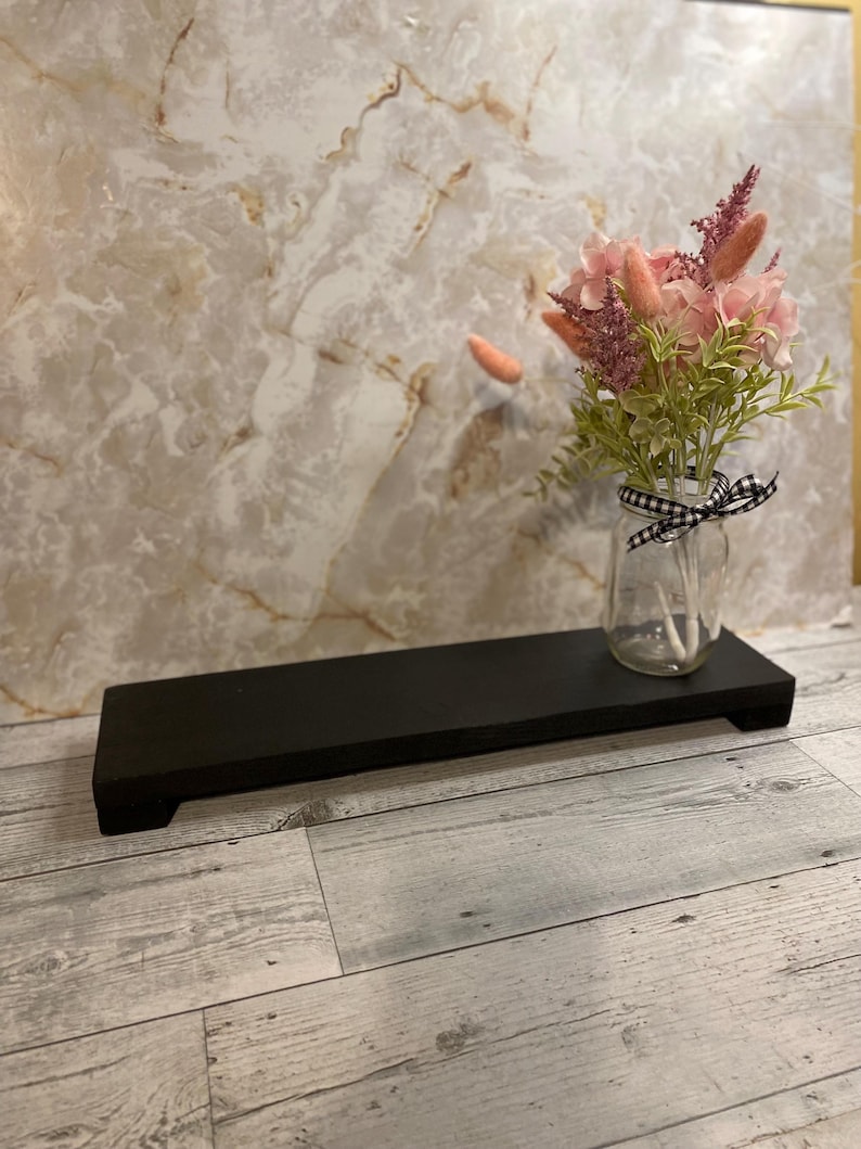 May include: A rectangular black wooden riser with short feet, featuring a clear glass jar filled with a bouquet of pink and green artificial flowers. The jar is decorated with a small black and white checkered ribbon tied around its neck.