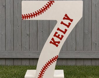 May include: A white, freestanding number seven decoration featuring red baseball-style stitching patterns across the top and bottom sections, with the name KELLY printed in bold red capital letters vertically along the center of the number.