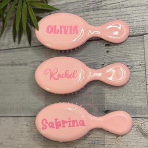 May include: Three pink, personalized hairbrushes. Each brush has a rounded oval head with a handle and bristles. The names "Olivia", "Rachel", and "Sabrina" are printed in pink script on each brush.