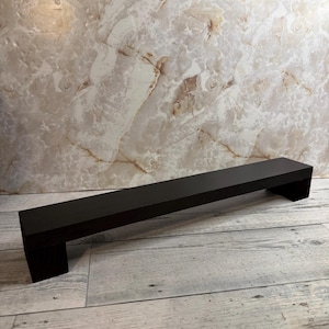 May include: A long, rectangular, matte black floating shelf with a solid, clean-lined construction. The shelf features a flat top surface and two vertical side supports that extend downward, creating a bridge-like structure.