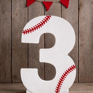 May include: A large white number three, designed to resemble a baseball. Red stitching details are on the number. A small string of red pennants hangs above the number. The background is a wooden wall.