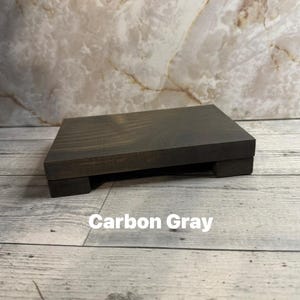 May include: A rectangular wooden riser with a dark carbon gray finish, featuring a flat top surface and four short, block-style feet. The text "Carbon Gray" is printed in white letters on the front of the wooden stand, which has a visible wood grain texture.