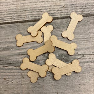 May include: A collection of small, wooden dog bone cutouts. The bones are light brown and have a classic bone shape with rounded ends. They are scattered on a light gray wooden surface.