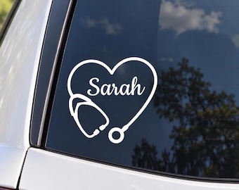 May include: A white vinyl decal applied to a car window featuring a heart-shaped outline formed by a stethoscope. The name Sarah is written in a white cursive script font inside the heart shape, centered within the stethoscope design.
