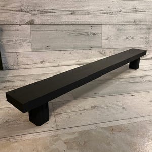 May include: A black wooden shelf with rounded edges and two square supports. The shelf is about 4 feet long and is resting on a white wood floor.