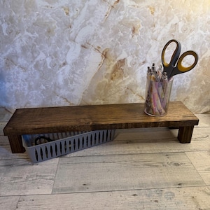 May include: A rectangular dark brown wooden riser shelf with four short legs. A clear plastic cup containing several pens and a pair of grey and yellow scissors sits on the right side. A grey plastic storage bin is tucked underneath the shelf.