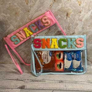 May include: Two clear zippered pouches with the word "SNACKS" in colorful, textured letters. One pouch is pink, the other is light blue, both with gold zippers and wrist straps. The blue pouch contains snack-sized treats.