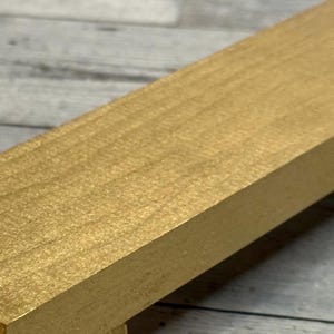 K&ouml;nnte beinhalten: Ein rechteckiger Holzblock mit einer glatten, metallisch goldenen Oberfl&auml;che. Das Objekt hat eine flache Oberseite und gerade, definierte Kanten, die die nat&uuml;rliche Maserung des Holzes unter der gleichm&auml;&szlig;igen Goldlackierung zeigen.