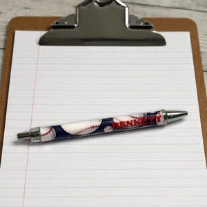 May include: A retractable ballpoint pen featuring a dark blue background with a white baseball pattern and red stitching details. The name BENNETT is printed in bold red capital letters along the center of the pen barrel, which has silver metal tips.