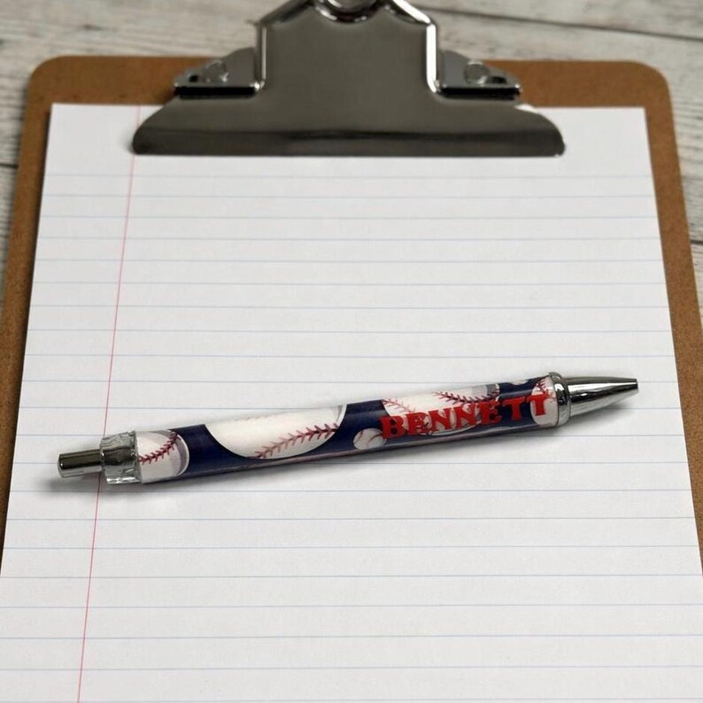 May include: A retractable ballpoint pen featuring a dark blue background with a white baseball pattern and red stitching details. The name BENNETT is printed in bold red capital letters along the center of the pen barrel, which has silver metal tips.