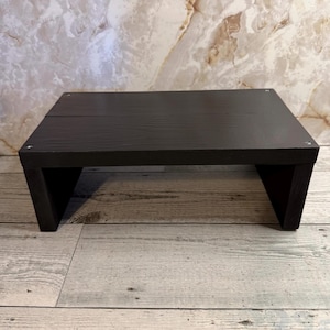 May include: A dark brown wooden step stool with a rectangular top and angled legs. The stool is made of wood and has a simple, modern design. The step stool is suitable for various uses.