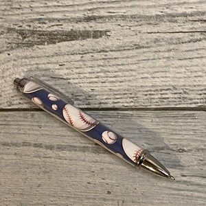 May include: A retractable ballpoint pen featuring a dark blue barrel decorated with a repeating pattern of white baseballs with red stitching. The pen includes a clear plastic pocket clip, a metallic silver tip, and a metallic silver plunger button.