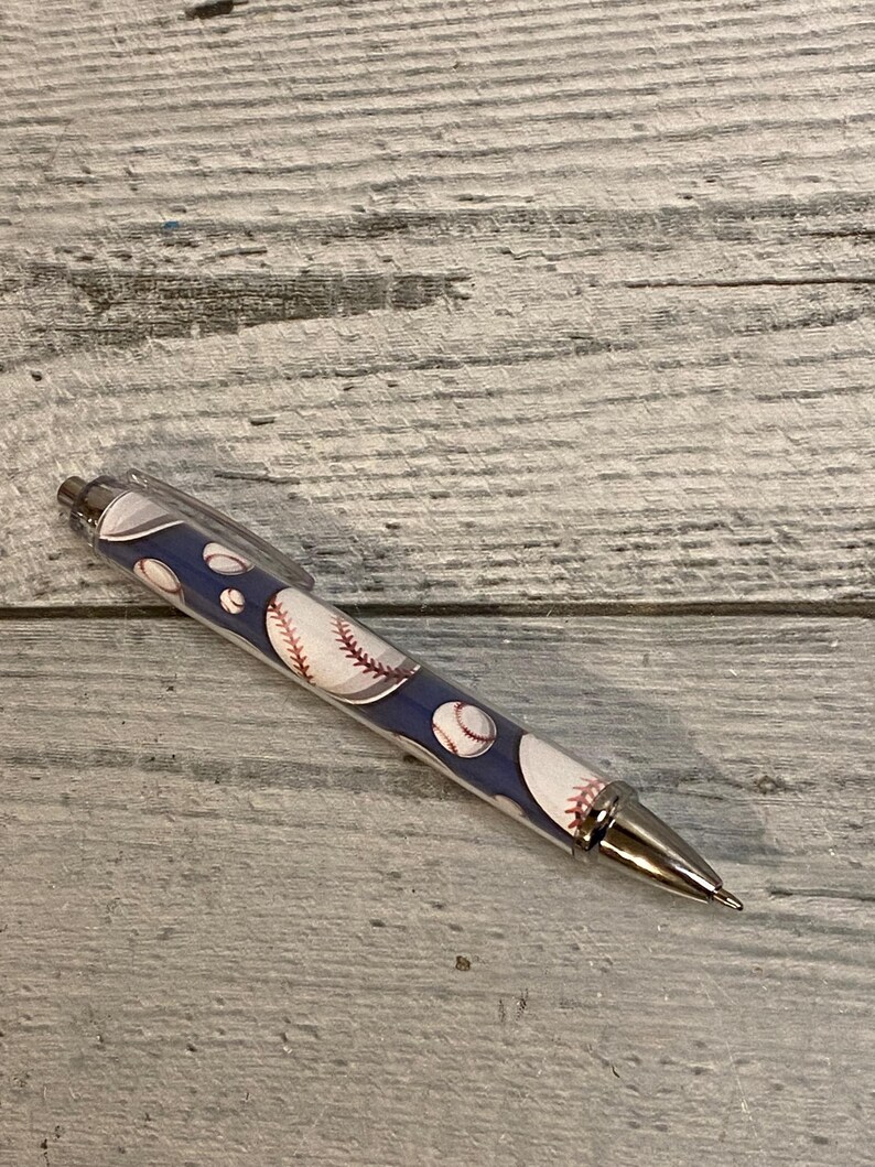 May include: A retractable ballpoint pen featuring a dark blue barrel decorated with a repeating pattern of white baseballs with red stitching. The pen includes a clear plastic pocket clip, a metallic silver tip, and a metallic silver plunger button.