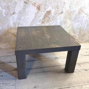 May include: A square wooden riser with a dark gray stained finish and a visible wood grain texture. The platform is supported by four thick, block-style legs and features four small, silver-colored metal screws visible on the top surface corners.
