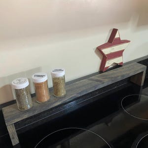 May include: A rustic wooden spice rack with three glass spice jars and a decorative star. The jars are labeled with spice names. The star is red and white. The rack is placed on a black surface.