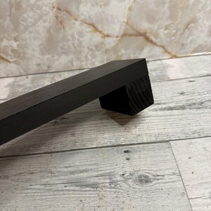 K&ouml;nnte beinhalten: Ein schwarzer M&ouml;belgriff aus Holz mit rechteckigem Profil und glatter, matter Oberfl&auml;che. Der Griff besteht aus einer langen, geraden Stange, die an einem quadratischen Montageblock befestigt ist und eine sichtbare Holzmaserung aufweist.