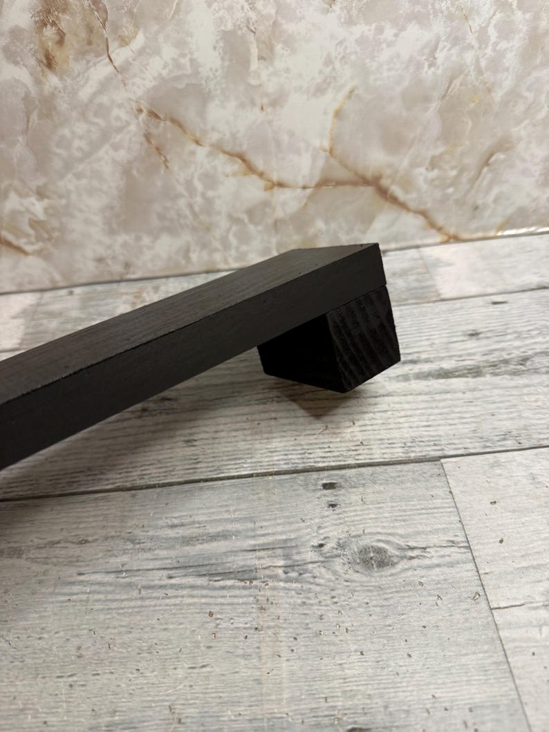 K&ouml;nnte beinhalten: Ein schwarzer M&ouml;belgriff aus Holz mit rechteckigem Profil und glatter, matter Oberfl&auml;che. Der Griff besteht aus einer langen, geraden Stange, die an einem quadratischen Montageblock befestigt ist und eine sichtbare Holzmaserung aufweist.