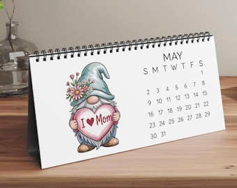 Könnte beinhalten: Ein weißer Tischkalender für den Monat Mai mit schwarzer Spiralbindung. Der Kalender zeigt eine Illustration eines Gnoms mit einer blauen Mütze, die mit rosa und weißen Blumen verziert ist und ein rosa Herz mit dem Text I Love Mom hält.