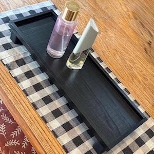 May include: A rectangular black wooden tray with short legs, holding a clear bottle with a gold cap containing pink liquid and a white bottle with a clear cap containing yellow liquid. The tray has a smooth finish and raised edges.