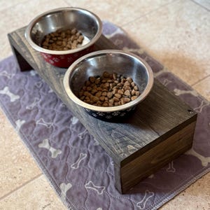 May include: A raised wooden dog bowl stand with two stainless steel bowls filled with dog food. One bowl has a red floral pattern, the other has a blue paw print design. The stand is on a purple bone-patterned mat.