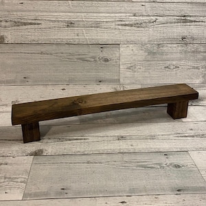 May include: A rectangular wooden riser with a dark brown stained finish, featuring a long flat top surface supported by two short, thick block legs at each end, showcasing a visible wood grain texture throughout the entire structure.