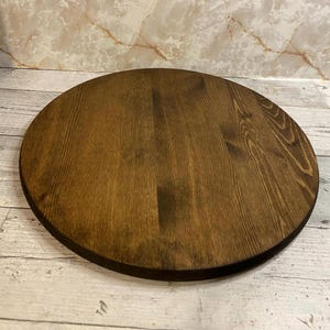 May include: A round, wooden serving tray with a dark brown finish. The tray has a smooth surface with visible wood grain and a slightly raised edge. The tray is suitable for serving food or as a decorative piece.