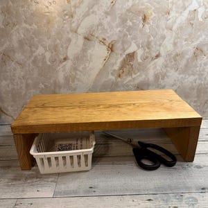 May include: A rectangular wooden desk riser with a natural finish. A small white basket is placed underneath the riser. A pair of black scissors rests on the surface. The desk riser is a home office accessory.