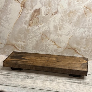 May include: A rectangular wooden riser with a dark brown stained finish and visible wood grain patterns. The platform is elevated by two small, dark-colored block feet positioned at each end, creating a low-profile display stand.