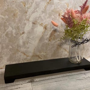 May include: A rectangular black wooden riser with short feet, featuring a clear glass jar tied with a black and white gingham ribbon. The jar contains a bouquet of pink and mauve dried flowers with green foliage, creating a floral arrangement.