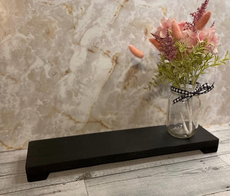 May include: A rectangular black wooden riser with short feet, featuring a clear glass jar tied with a black and white gingham ribbon. The jar contains a bouquet of pink and mauve dried flowers with green foliage, creating a floral arrangement.