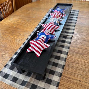 May include: A black wooden tray with six star-shaped patriotic decorations. The stars feature red and white stripes and blue backgrounds with white stars. The tray sits on a black and white checkered runner.