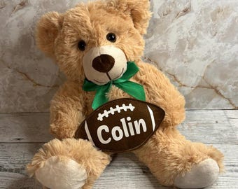 Könnte beinhalten: Ein hellbrauner Plüschteddybär mit dunklen Augen und einer braunen Nase. Der Bär trägt ein grünes Satinband um den Hals und hält einen braunen, fußballförmigen Filzaufnäher mit weißen Nähten und dem Namen Colin in weißer Schrift.