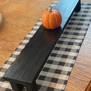 May include: A long, rectangular black wooden riser with four short legs, featuring a smooth, dark finish. A small, round, orange decorative pumpkin with a brown stem is positioned on the center of the flat surface of the wooden riser.