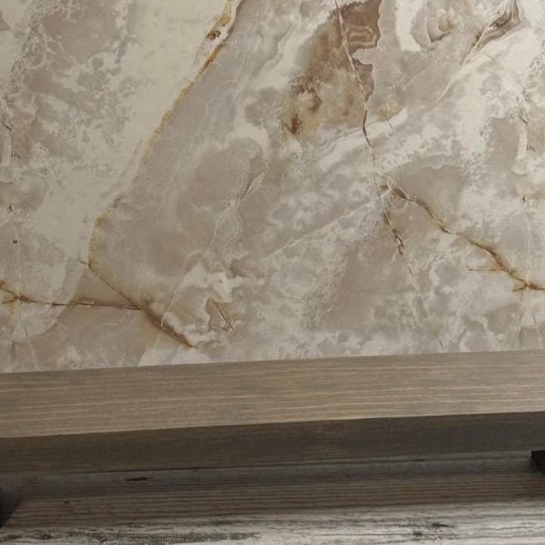 K&ouml;nnte beinhalten: Ein horizontales Holzregal mit einer glatten, mittelbraunen Oberfl&auml;che ist vor einer gro&szlig;en, polierten Platte aus beigem und cremefarbenem Stein positioniert, die durchgehend markante, flie&szlig;ende braune Maserungen aufweist.