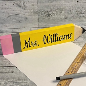 May include: A wooden pencil-shaped desk sign painted yellow with a pink eraser, a grey ferrule, and a black tip. The text "Mrs. Williams" is printed in black script on the yellow body. A wooden ruler is visible, showing a length of 9 inches.