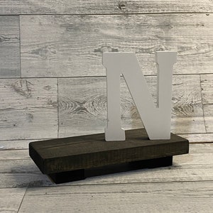 May include: White wooden letter N on a dark brown wooden base. The base is made of reclaimed wood and has a rustic look.