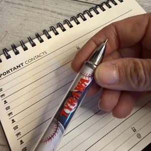 May include: A ballpoint pen featuring a baseball pattern and the name TYRAWA printed in red letters. The pen has a silver-toned metal tip and a clear barrel, positioned over a spiral-bound notebook with lined pages labeled for important contacts.