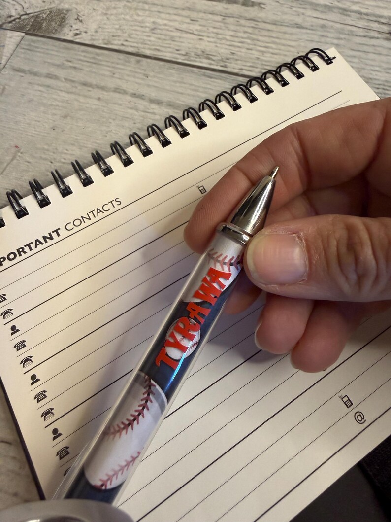 May include: A ballpoint pen featuring a baseball pattern and the name TYRAWA printed in red letters. The pen has a silver-toned metal tip and a clear barrel, positioned over a spiral-bound notebook with lined pages labeled for important contacts.