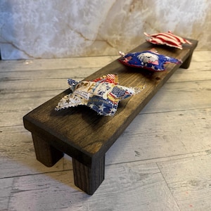 May include: A rectangular dark brown wooden riser with four short legs, featuring three fabric star-shaped ornaments on top. The stars display patriotic patterns including red, white, and blue colors, American flag motifs, and text that reads "One Nation Under God, Indivisible, We Still Stand."