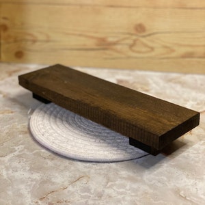 May include: A dark brown wooden shelf with black supports. The shelf is rectangular and has a natural wood grain. It is placed on a white, circular, woven coaster. The background is a light brown wooden surface.