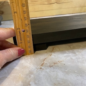 May include: A dark brown wooden riser or shelf with a rectangular shape. A wooden ruler is positioned vertically against the side of the object, showing a thickness of approximately 1 inch. The wood has a visible grain and a smooth, dark finish.