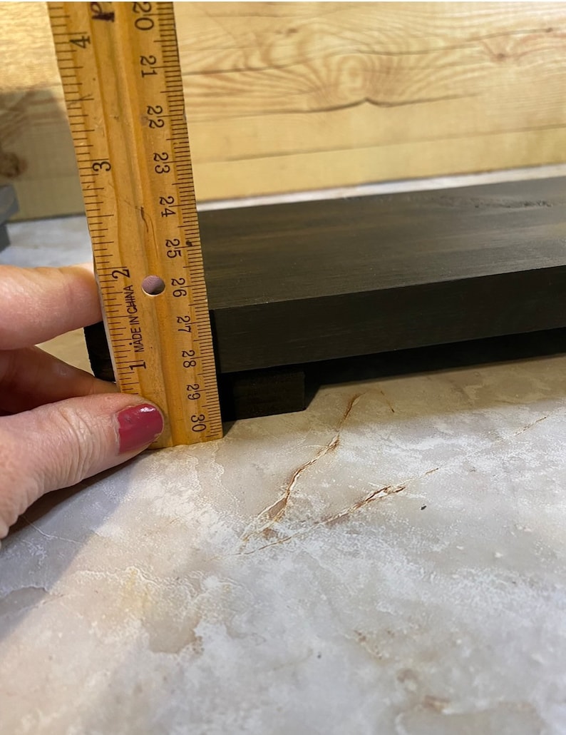 May include: A dark brown wooden riser or shelf with a rectangular shape. A wooden ruler is positioned vertically against the side of the object, showing a thickness of approximately 1 inch. The wood has a visible grain and a smooth, dark finish.